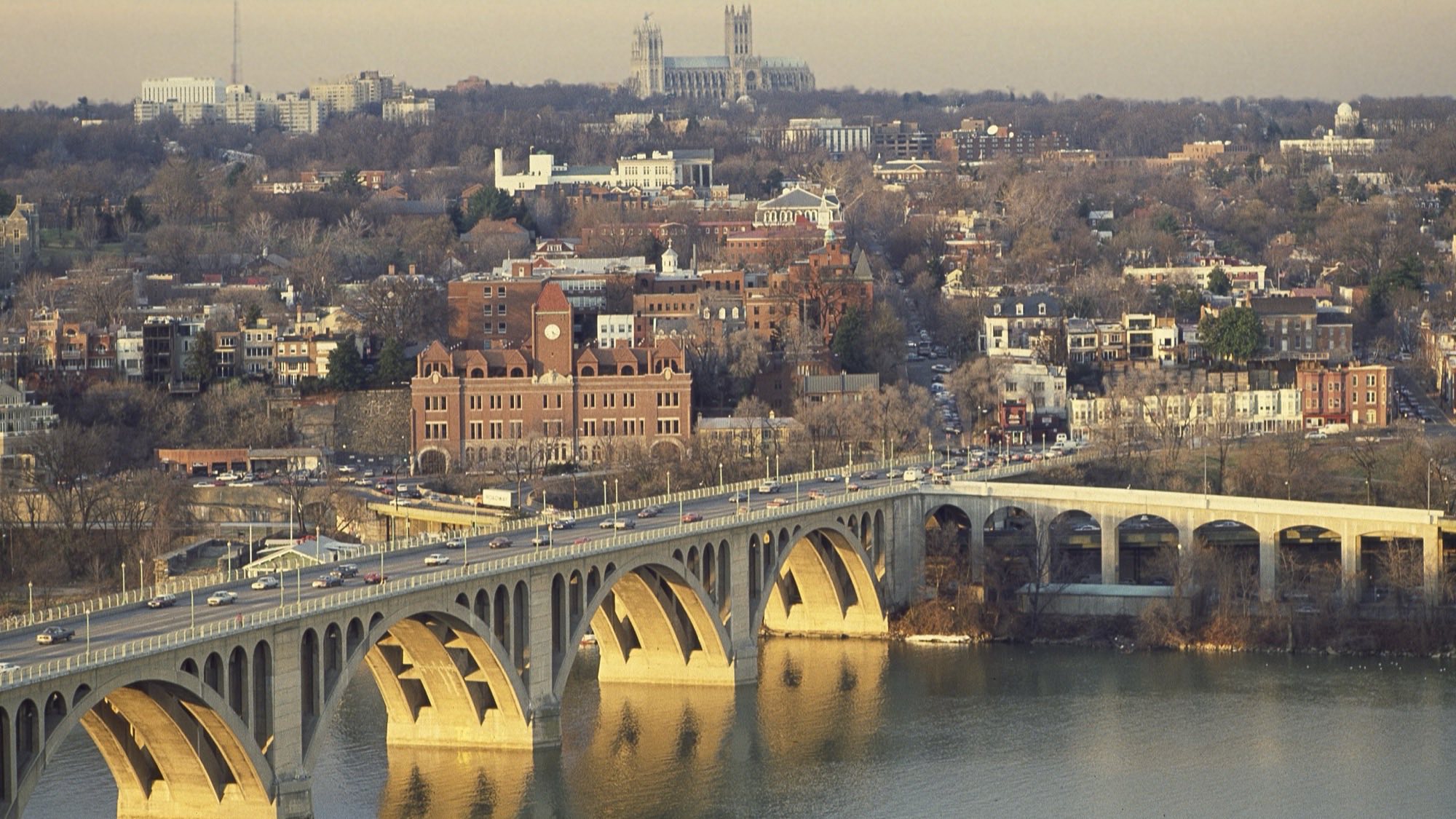 Key Bridge over the Potomac at golden hour, Georgetown rowhouses in the mid-ground and Washington National Cathedral on the ridgeline beyond -- shot from the Ro