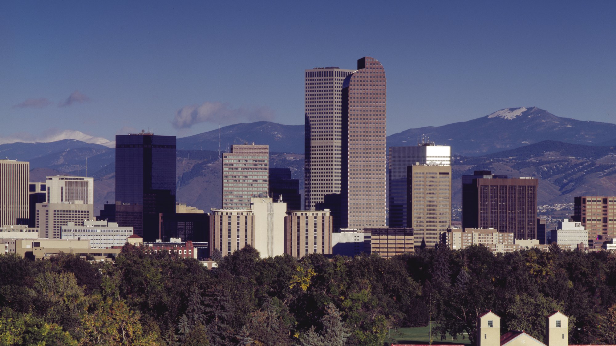 Downtown Denver skyline (1801 California’s rounded crown dominant) framed by the snowcapped Front Range on both flanks, City Park treeline in the foreground --