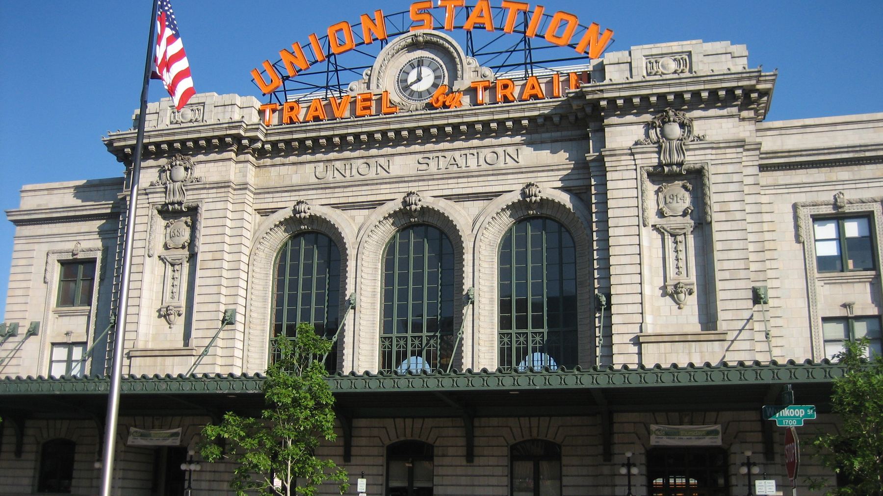 Denver Union Station’s 1914 facade with the lit orange "UNION STATION / TRAVEL BY TRAIN" sign and clock tower, American flag, clear warm morning light