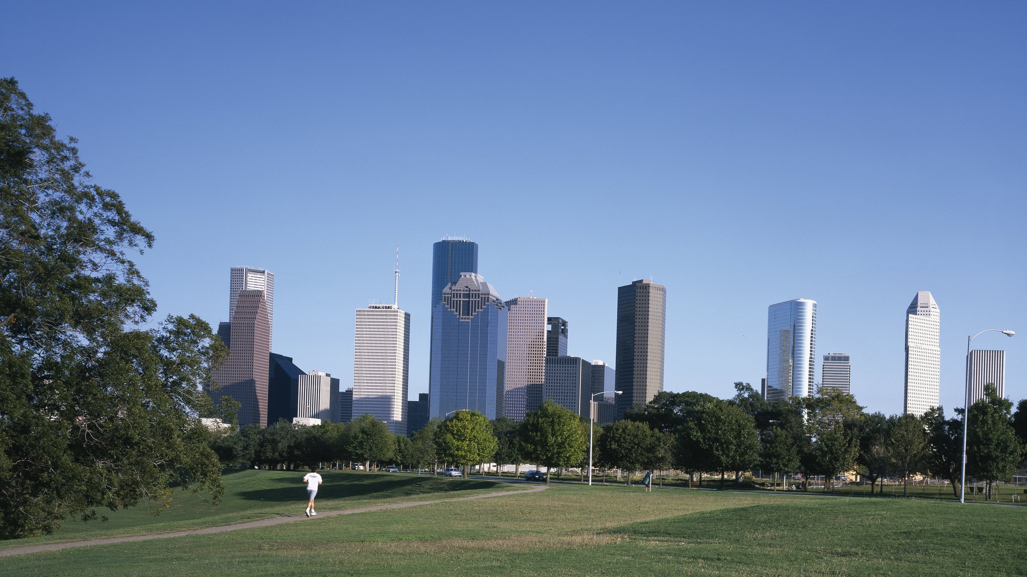 Downtown Houston skyline seen from Buffalo Bayou Park, jogger on the path, live-oak tree canopy foreground -- brief VISUAL IDENTITY #1 subject
