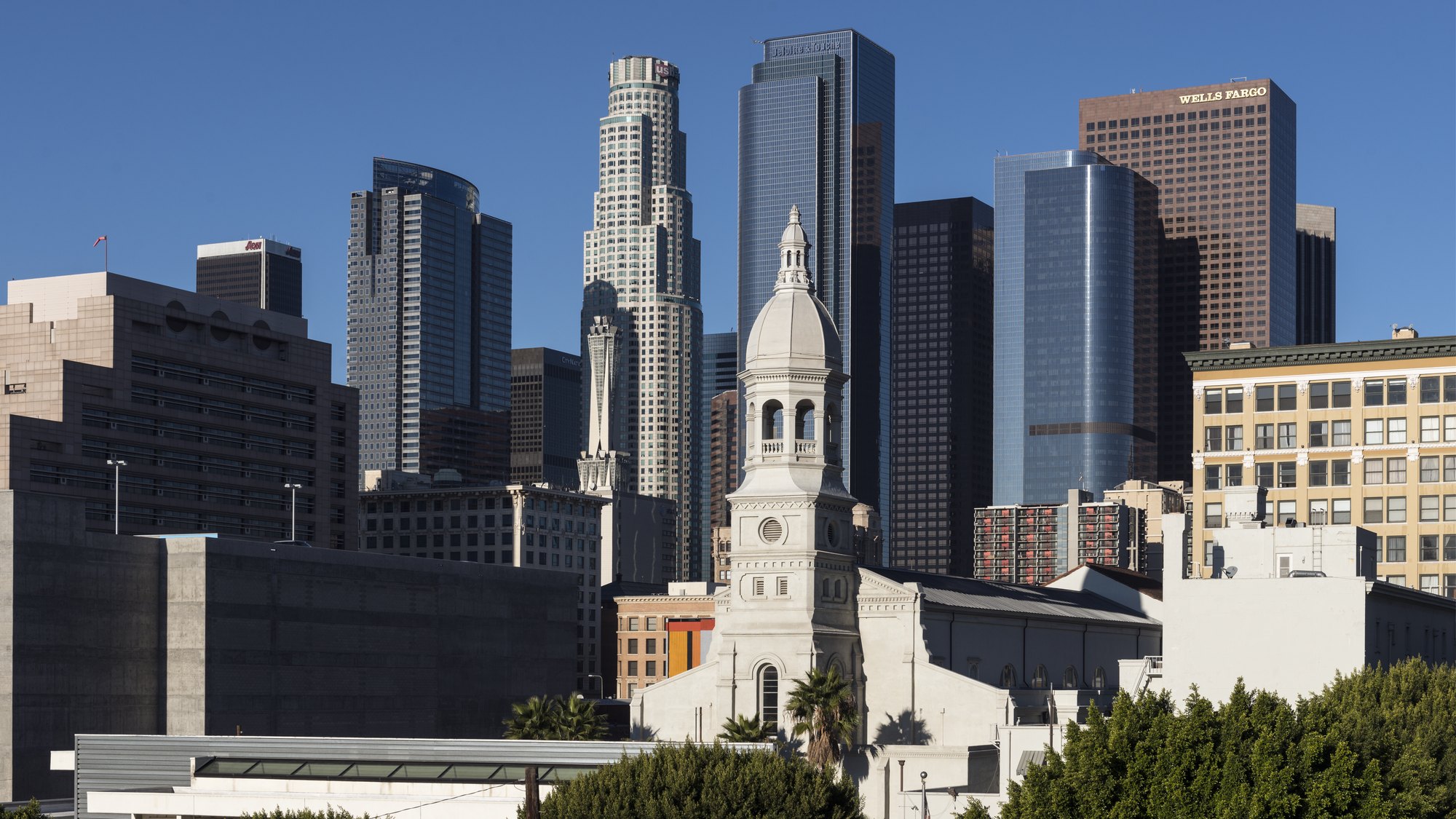 Downtown Los Angeles skyline (US Bank Tower, Wells Fargo Tower, Deloitte tower cluster) with St. Vibiana Cathedral bell tower and palm trees in foreground, clea