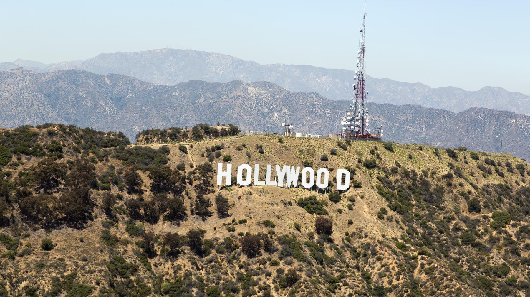 The Hollywood Sign, aerial oblique from Mount Lee, San Gabriel Mountains layered behind in haze, KTLA/broadcast tower on the ridgeline, clear daylight -- brief