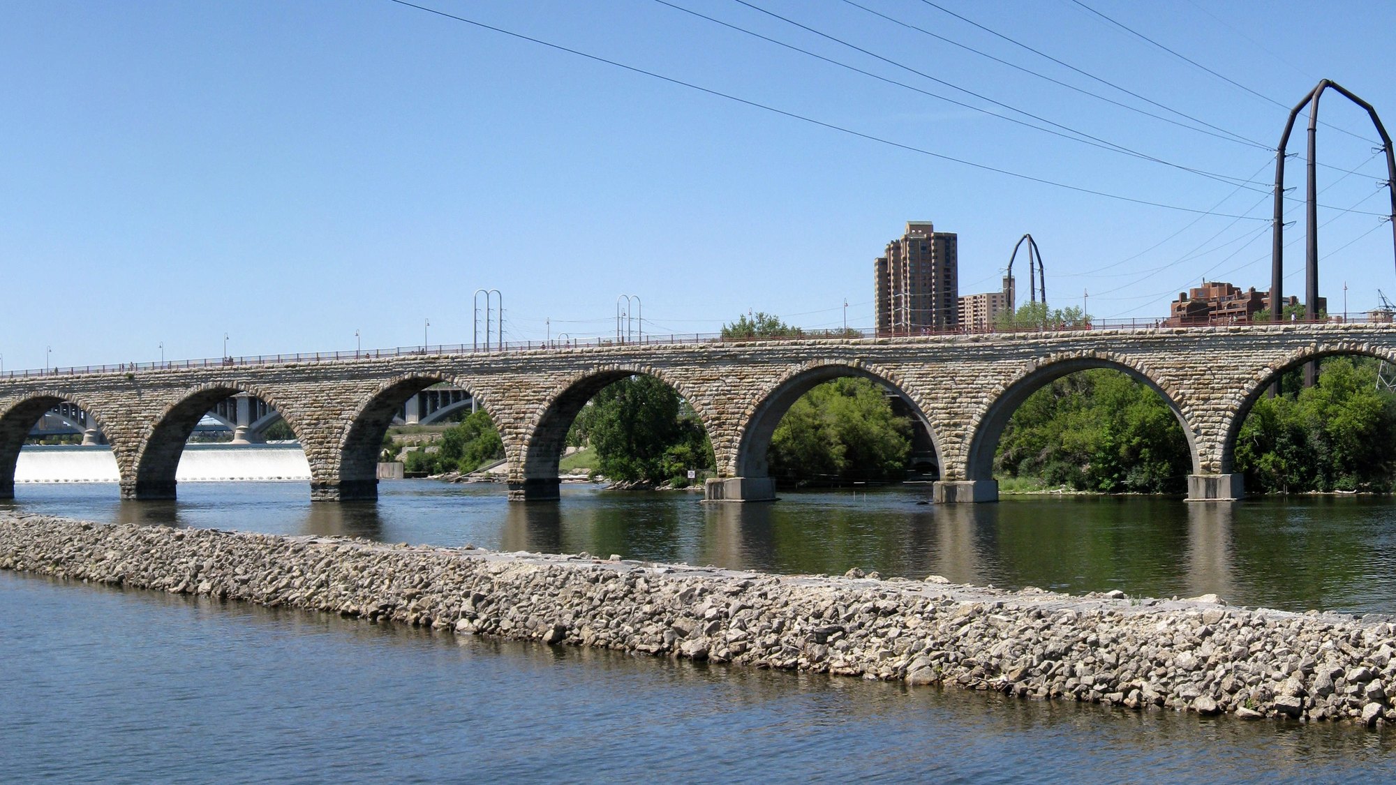Stone Arch Bridge full seven-arch span over St. Anthony Falls spillway, Mississippi River rocky bank foreground, Minneapolis downtown skyline (including the Gol