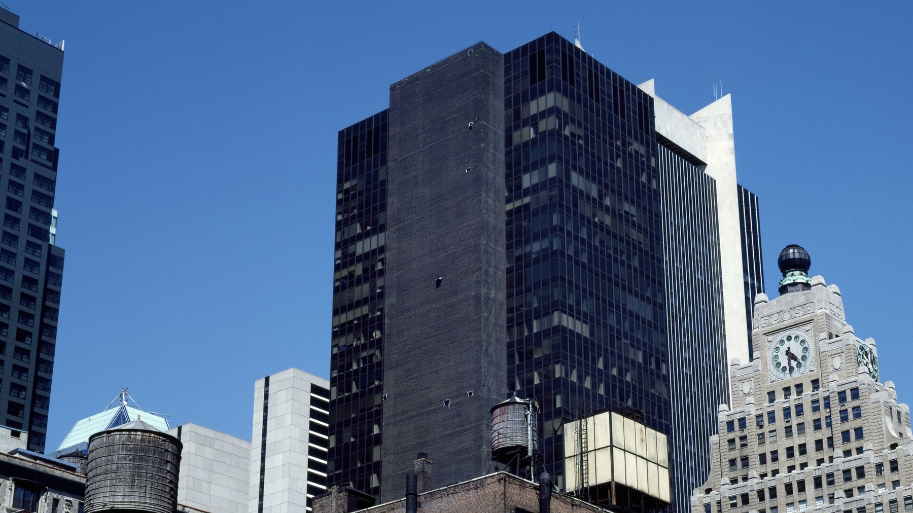 Wooden rooftop water tower on a Manhattan tenement building, downtown skyline (ornate clock-tower building) behind, clear blue daylight -- the resident’s skylin