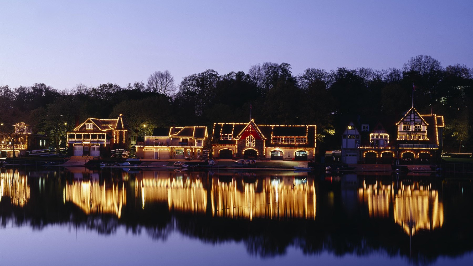 Boathouse Row at dusk, Kelly Drive on the Schuylkill River -- full Victorian boathouse row (Vesper and Malta name-boards legible) traced edge-to-edge in warm st