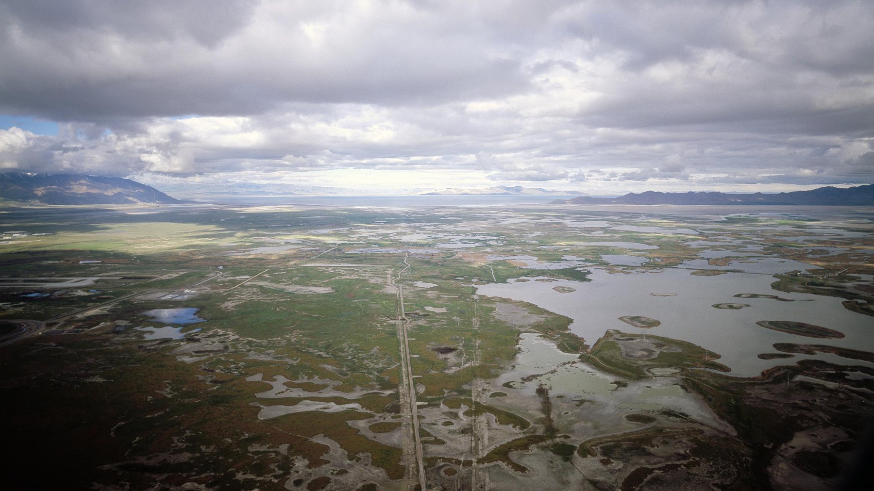 Eastern edge of the Great Salt Lake -- elevated aerial over the salt-crusted wetlands and causeway road, open lake water and the Wasatch/Oquirrh ranges layered