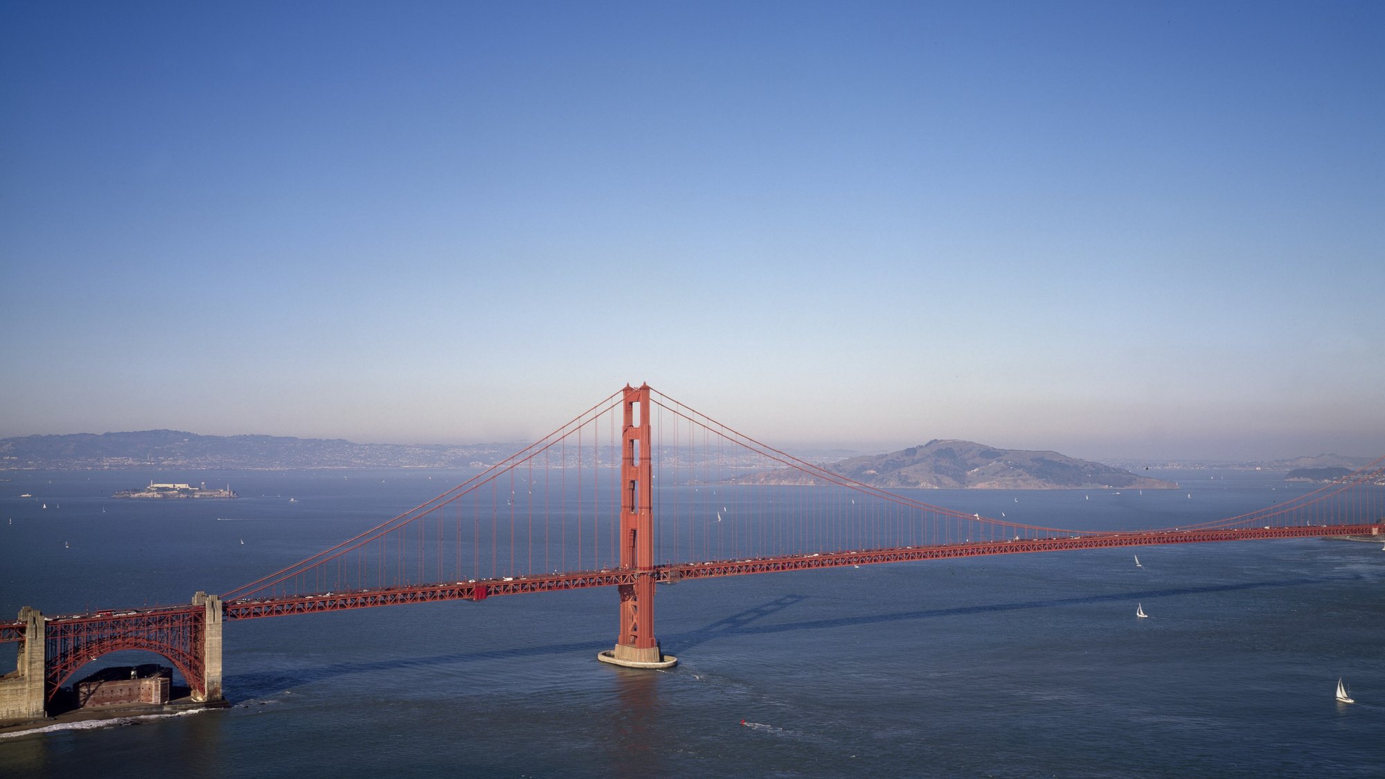 Golden Gate Bridge full tower, elevated oblique aerial view from the south side of the strait at golden-hour, warm pink-orange sky glow, full suspension cables